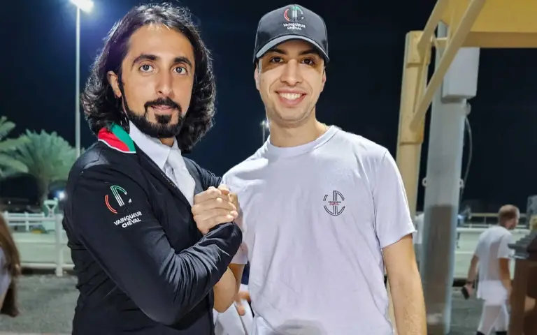 Two men shaking hands, one wearing a black jacket and the other in a white t-shirt and cap, both showcasing VC Wear apparel.