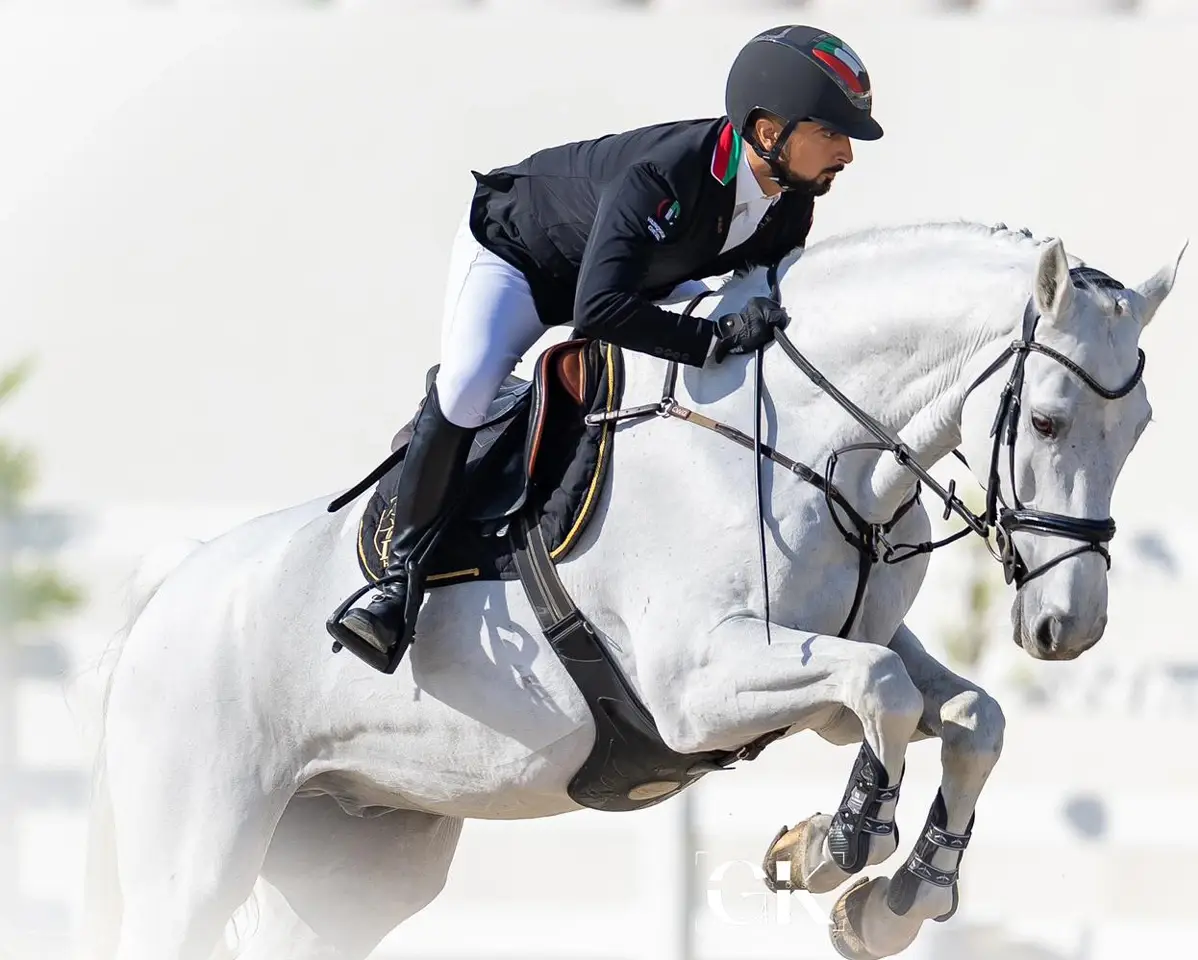 Equestrian rider in a black jacket and white pants jumping over a hurdle on a white horse, showcasing VC Wear.